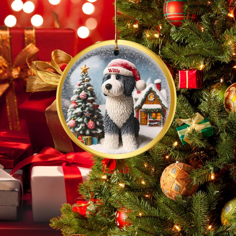 May include: A circular Christmas decoration with a gold edge depicts a dog in a Santa hat. The ornament shows a winter scene with a Christmas tree and a snow-covered house. The dog's name, "Willis", is on the hat.