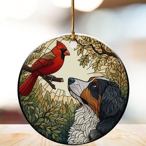 May include: Round ceramic ornament featuring a red cardinal perched on a branch, facing a Bernese Mountain Dog. The dog has black, white, and brown fur. The background includes trees and a light-colored sky, with a gold string for hanging.