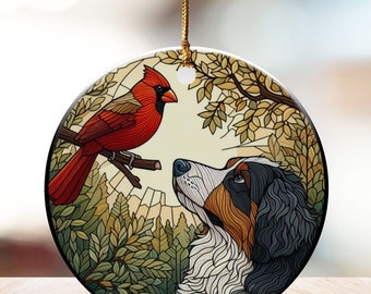May include: Round ceramic ornament featuring a red cardinal perched on a branch, facing a Bernese Mountain Dog. The dog has black, white, and brown fur. The background includes trees and a light-colored sky, with a gold string for hanging.