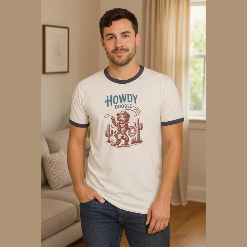 May include: Cream-colored ringer t-shirt with navy blue trim featuring a cartoon cowboy dog graphic and the words "Howdy Doodle". The dog is holding a lasso, with cacti on either side.