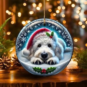 Puede incluir: Adorno navideño redondo con un perro blanco con un gorro de Papá Noel rojo con la palabra "Snowflake". El adorno tiene un fondo nevado con copos de nieve y acentos de acebo. Se adjunta una cuerda dorada para colgarlo.
