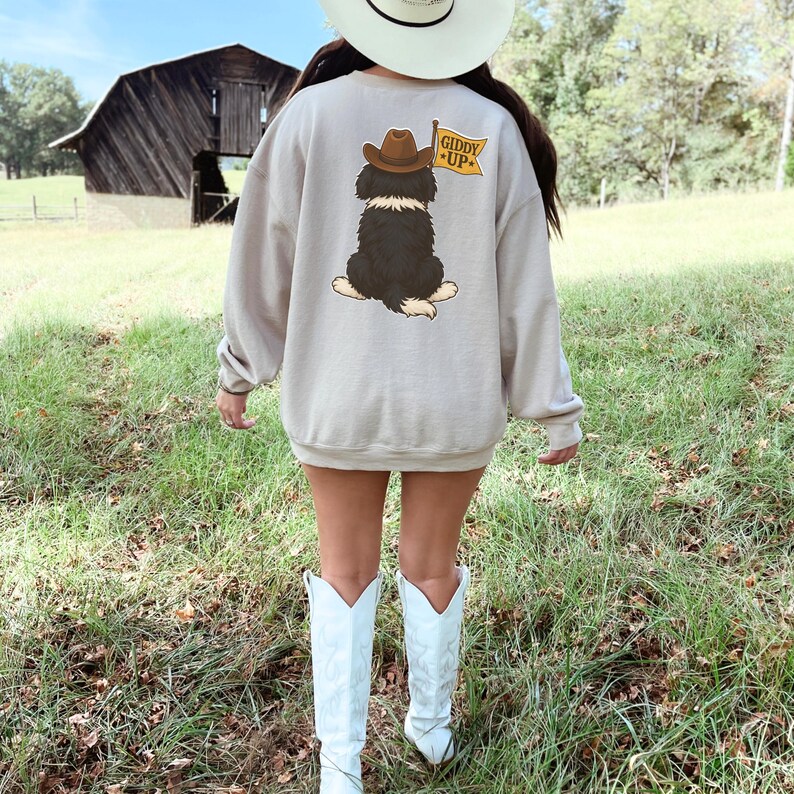 Puede incluir: Sudadera beige con un dibujo de un perro con sombrero de vaquero y una bandera con la inscripci&oacute;n "Giddy Up". El conjunto incluye botas de vaquero blancas y un sombrero de vaquero blanco. El fondo muestra un granero y hierba verde.