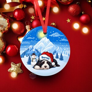 May include: A round Christmas ornament featuring a black and white dog wearing a Santa hat with the name "Bernie". The ornament has a winter scene with a snowman, mountains, and a blue sky. A red ribbon is attached for hanging.