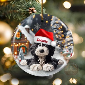 May include: A round Christmas ornament featuring a black and white dog wearing a red Santa hat with the name "Zoomie". The ornament has a winter scene background with a house and falling snow. A gold string is attached for hanging.