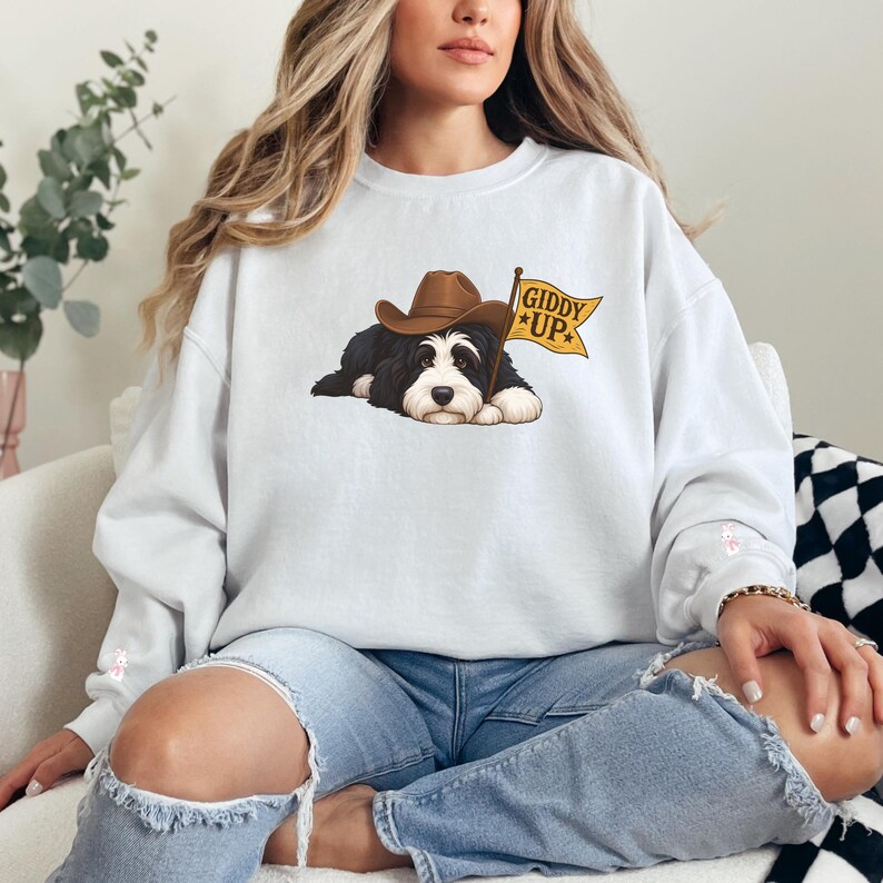Puede incluir: Sudadera blanca con un dise&ntilde;o de un perro blanco y negro con un sombrero de vaquero marr&oacute;n. El perro est&aacute; tumbado junto a una bandera amarilla con la inscripci&oacute;n "Giddy Up".