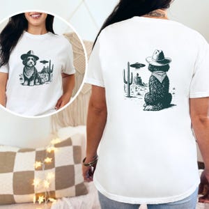 May include: White t-shirt featuring a black and white graphic of a dog wearing a cowboy hat and bandana, with a UFO and desert landscape. The design is on the front and back of the shirt.