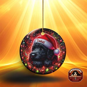 May include: A round Christmas ornament featuring a black dog wearing a Santa hat with the name "Teddi" on it. The ornament has a festive design with red and green accents, and a gold string for hanging.
