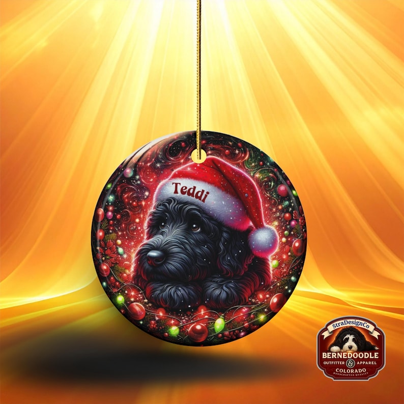 May include: A round Christmas ornament featuring a black dog wearing a Santa hat with the name "Teddi" on it. The ornament has a festive design with red and green accents, and a gold string for hanging.