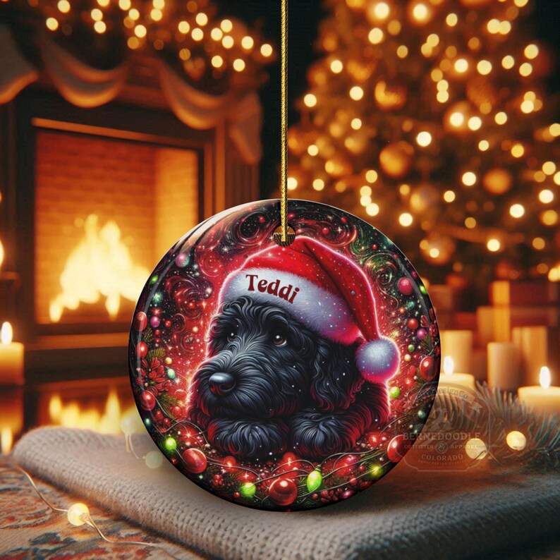 May include: A round Christmas ornament featuring a black dog wearing a Santa hat with the name "Teddi". The ornament is decorated with festive lights and a red background. A gold string is attached for hanging.