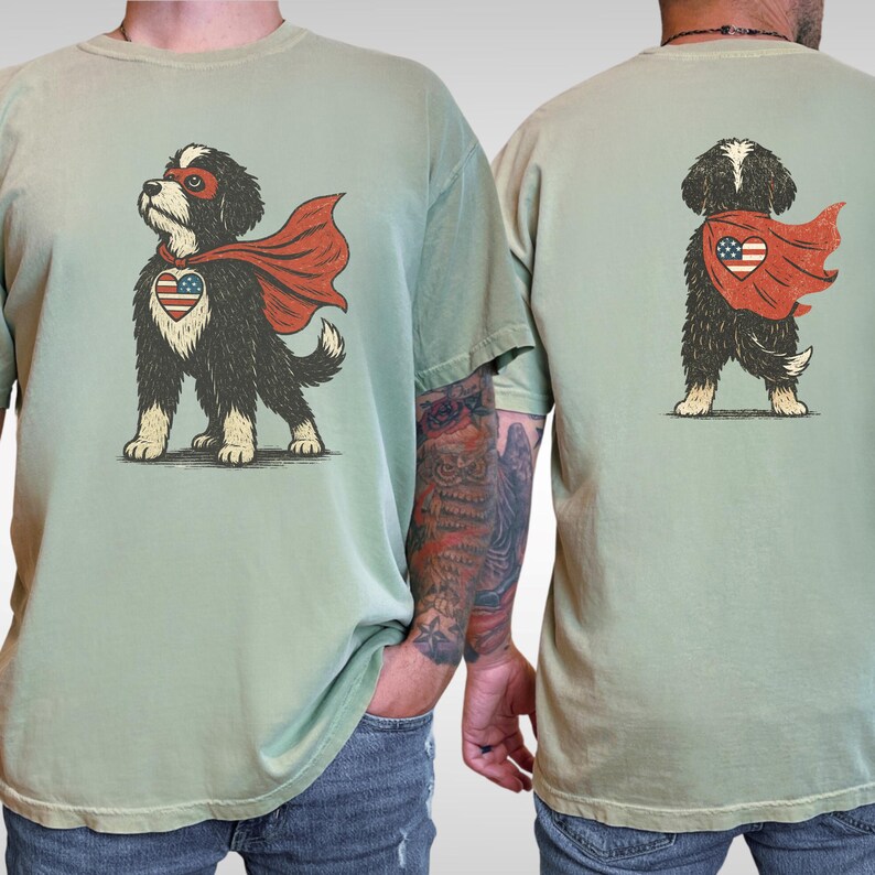 May include: A sage green t-shirt featuring a graphic of a black and white dog wearing a red superhero cape and a red eye mask. The dog has a heart-shaped patch on its chest displaying the American flag. The same graphic appears on the back.