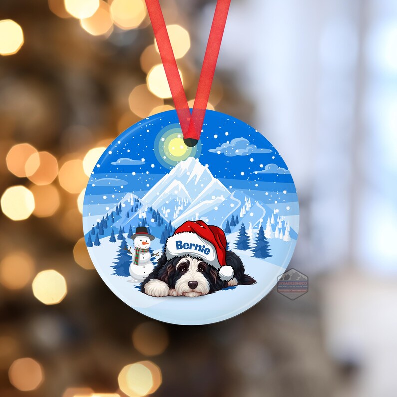 May include: Round Christmas ornament featuring a Bernese Mountain Dog wearing a Santa hat with the name "Bernie". The ornament has a winter scene with a snowman, mountains, and a blue sky. A red ribbon is attached for hanging.