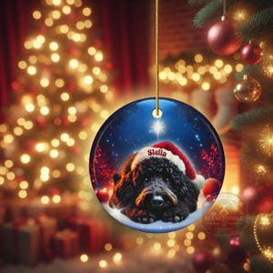 May include: A round Christmas ornament featuring a black dog wearing a Santa hat with the name "Stella". The ornament has a blue background with a star and snow. The ornament is hanging from a gold string.