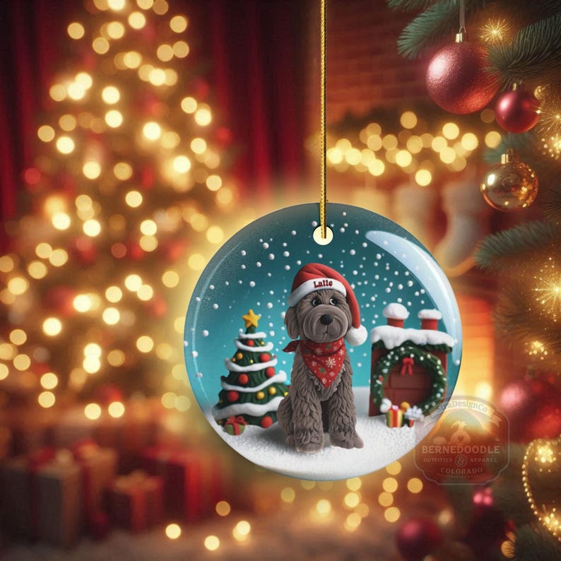 May include: A round Christmas ornament featuring a brown dog wearing a Santa hat and a red bandana. The ornament has a snowy scene with a Christmas tree and a house. The dog's name, "Lalle", is on the hat.