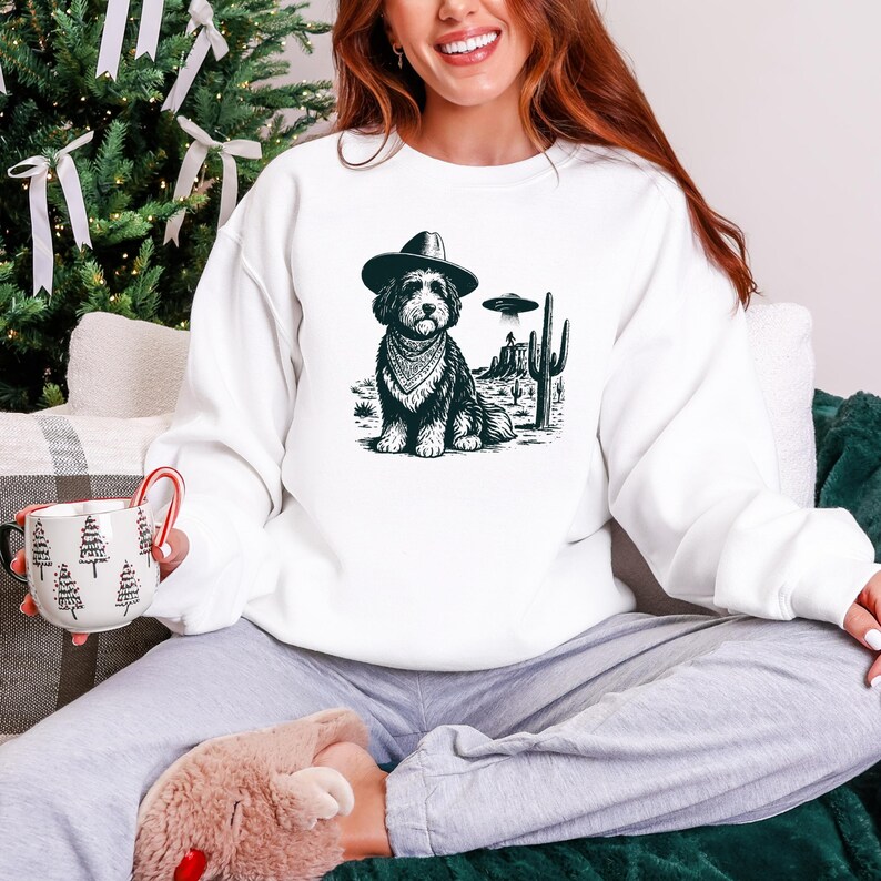 May include: White sweatshirt featuring a black and white graphic of a dog wearing a cowboy hat and bandana, set against a desert landscape with a UFO. The dog is the central focus, with cacti and a flying saucer adding detail.
