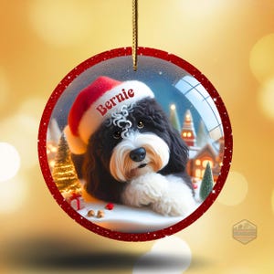 K&ouml;nnte beinhalten: Runder Weihnachtsschmuck mit einem schwarz-wei&szlig;en Hund, der einen Weihnachtsmannhut mit dem Namen "Bernie" tr&auml;gt. Der Schmuck hat einen roten Glitzerrahmen und eine goldene Schnur zum Aufh&auml;ngen. Der Hintergrund zeigt eine Winterlandschaft mit Schnee und einem geschm&uuml;ckten Weihnachtsbaum.