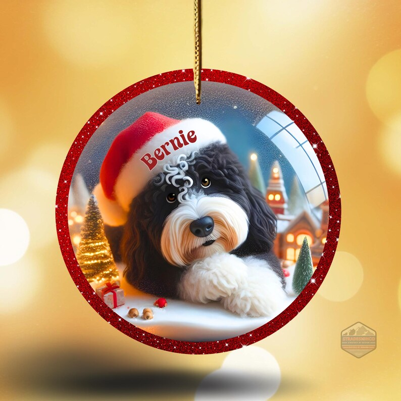 K&ouml;nnte beinhalten: Runder Weihnachtsschmuck mit einem schwarz-wei&szlig;en Hund, der einen Weihnachtsmannhut mit dem Namen "Bernie" tr&auml;gt. Der Schmuck hat einen roten Glitzerrahmen und eine goldene Schnur zum Aufh&auml;ngen. Der Hintergrund zeigt eine Winterlandschaft mit Schnee und einem geschm&uuml;ckten Weihnachtsbaum.