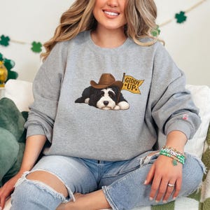 Puede incluir: Una sudadera gris jaspeada con un gr&aacute;fico de un perro blanco y negro con un sombrero de vaquero marr&oacute;n. El perro est&aacute; acostado junto a una bandera amarilla que dice "Giddy Up". La sudadera es de cuello redondo.