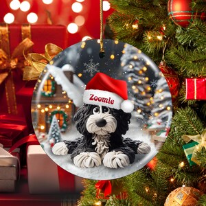 May include: A round Christmas ornament featuring a black and white dog wearing a red Santa hat with the name "Zoomie". The ornament has a winter scene background with a house and snow. The ornament is hanging on a Christmas tree.