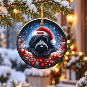 May include: A round ceramic Christmas ornament featuring a black dog wearing a red Santa hat with the name Stella printed on the brim. The dog is surrounded by red berries and ornaments against a dark blue snowy background with a gold string.