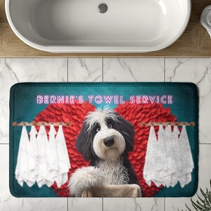 May include: A rectangular bath mat featuring a cartoon dog with black and white fur, standing in front of a red heart-shaped design. The mat has the text "BERNIE'S TOWEL SERVICE" in pink neon lettering. White towels hang on a wooden rod.