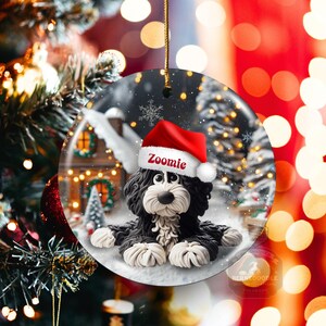 May include: A round Christmas ornament featuring a black and white dog wearing a red Santa hat with the name "Zoomie". The ornament has a winter scene background with a house and snow. A gold string is attached for hanging.