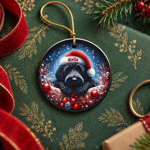 May include: A round ceramic Christmas ornament featuring a black dog wearing a red and white Santa hat with the name Stella printed on the brim. The dog is surrounded by red berries, sparkling lights, and a snowy blue background.