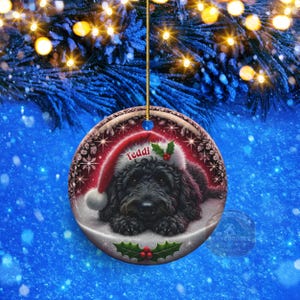 May include: A round Christmas ornament featuring a black dog wearing a Santa hat with the name "Teddi". The ornament has a red and white border with snowflakes and holly accents. It hangs from a gold string against a blue background.