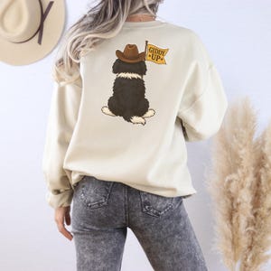 Puede incluir: Sudadera color crema con un perro de dibujos animados con un sombrero de vaquero y una bandera que dice "Giddy Up". El perro es blanco y negro con un sombrero marr&oacute;n y una bandera amarilla. La sudadera es de color neutro.