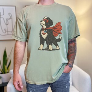 May include: A sage green t-shirt featuring a graphic illustration of a black and white dog wearing a red superhero mask and cape. The dog has a heart-shaped patch on its chest with a red, white, and blue American flag pattern.