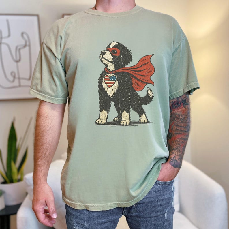 May include: A sage green t-shirt featuring a graphic illustration of a black and white dog wearing a red superhero mask and cape. The dog has a heart-shaped patch on its chest with a red, white, and blue American flag pattern.