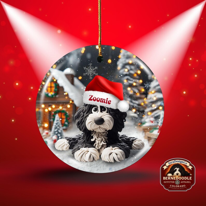 May include: A round Christmas ornament featuring a Bernedoodle dog wearing a Santa hat with the name "Zoomie". The ornament has a snowy winter scene background with a house and Christmas tree. A gold string is attached for hanging.