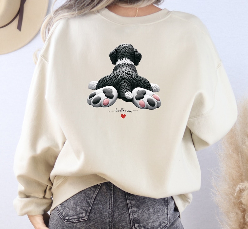 Unisex crewneck sweatshirt featuring an adorable claymation-style Bernedoodle dog illustration on the front, holding a red heart tag that can be personalized with a pet’s name. The back of the sweatshirt shows a cute rear-view Bernedoodle lying flat with detailed fluffy fur and oversized pink-paw pads, creating a playful “doodle mom” design. Ideal for Bernedoodle lovers, dog moms. Great for holidays, Mother’s Day, personalized pet gifts, and everyday cozy wear.