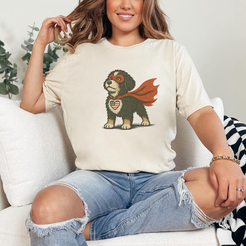 May include: Cream-colored t-shirt featuring a cartoon dog dressed as a superhero. The dog has a red cape, mask, and a heart-shaped emblem with the American flag design. The shirt is a casual style.