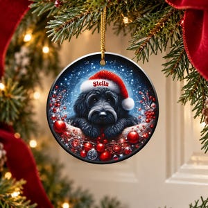 May include: A round ceramic Christmas ornament featuring a black dog wearing a red Santa hat with the name Stella printed on the brim. The dog is surrounded by red berries and ornaments against a dark blue, snowy background.