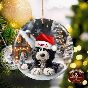 May include: A round Christmas ornament featuring a black and white Bernedoodle dog wearing a red Santa hat with the name "Zoomie". The ornament has a winter scene background with a snowy house and trees. A gold string is attached for hanging.