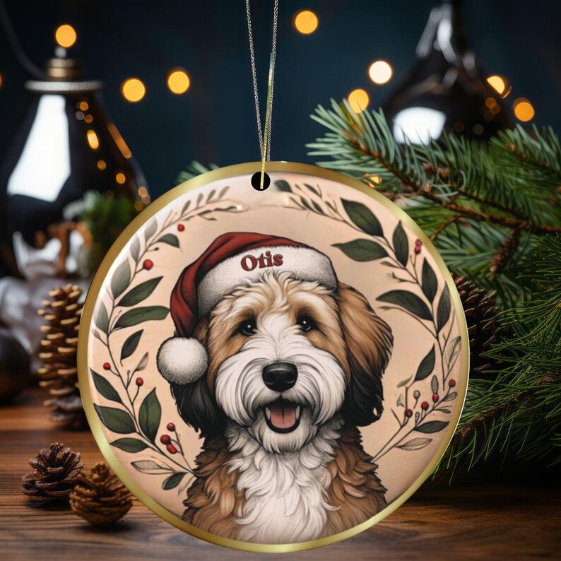 May include: Round Christmas ornament featuring a smiling dog wearing a Santa hat with the name "Otis". The ornament has a gold trim and is decorated with a wreath of green leaves and red berries. A festive holiday decoration.