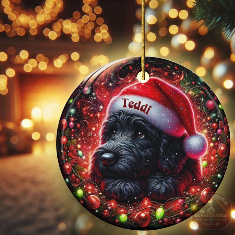 May include: A round Christmas ornament featuring a black dog wearing a Santa hat with the name "Teddi" on it. The ornament has a red and black background with Christmas lights and ornaments. A gold string is attached for hanging.