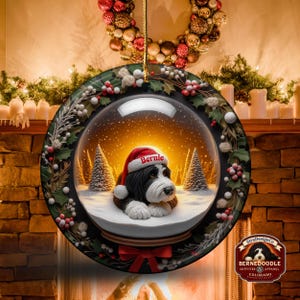 May include: A round Christmas ornament featuring a Bernedoodle dog inside a snow globe. The dog wears a Santa hat with the name "Bernie." The ornament is decorated with holly and berries, and a red bow. The background shows a fireplace.
