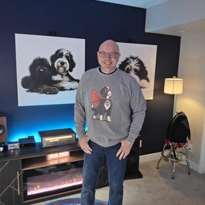 May include: A grey long-sleeve sweatshirt featuring a graphic of a dog wearing a red superhero cape and a heart-shaped patch with a star-spangled banner design on its chest, paired with dark blue denim jeans and white trainers with red stripes.