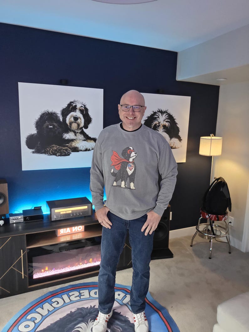 May include: A grey long-sleeve sweatshirt featuring a graphic of a dog wearing a red superhero cape and a heart-shaped patch with a star-spangled banner design on its chest, paired with dark blue denim jeans and white trainers with red stripes.