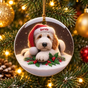 May include: A round white ceramic Christmas ornament featuring a fluffy cream and tan dog wearing a red Santa hat with the name Bernie printed on the brim. The dog rests on a snowy surface decorated with green holly leaves and red berries.
