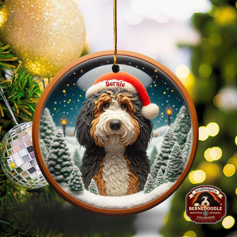Op de afbeelding: Een ronde kerstversiering met een Bernedoodle-hond met een kerstmuts met de naam "Bernie". De versiering heeft een houten frame en toont een besneeuwde wintersc&egrave;ne met dennenbomen en straatlantaarns. Het hangt aan een gouden touwtje.