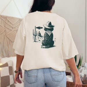 May include: Cream-colored t-shirt featuring a graphic of a dog wearing a cowboy hat and bandana, with a desert landscape and UFO. The dog is facing away from the viewer. The design is in shades of green and black.