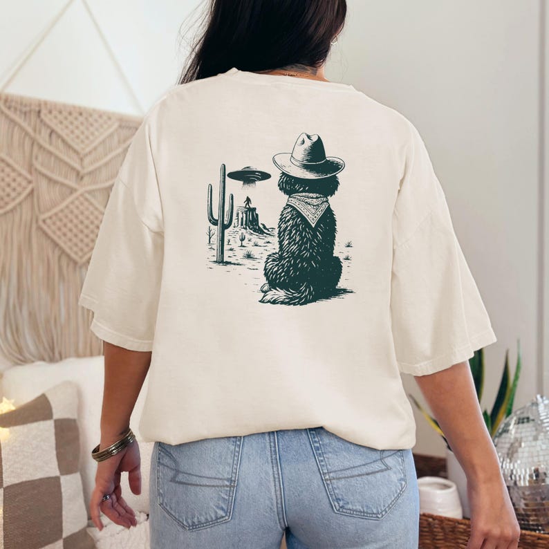 May include: Cream-colored t-shirt featuring a graphic of a dog wearing a cowboy hat and bandana, with a desert landscape and UFO. The dog is facing away from the viewer. The design is in shades of green and black.