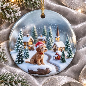 May include: A round ceramic Christmas ornament featuring a golden-brown dog wearing a red Santa hat with the name Goldie printed on the brim. The scene depicts a snowy village with pine trees, small houses, and a church under a light snowfall.