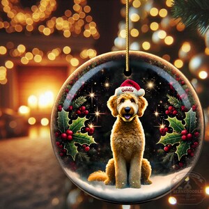 May include: A round Christmas ornament featuring a golden doodle dog wearing a Santa hat with the name "Goldie". The ornament is decorated with holly and berries, set against a snowy background with a starry night sky.