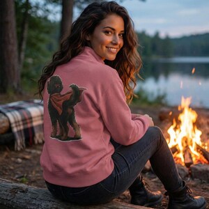 Pu&ograve; includere: Una felpa color rosa polveroso con una grafica di un cane nero e marrone con un mantello rosso. La felpa ha un colletto alto e una cerniera. La persona &egrave; seduta su un tronco vicino a un fal&ograve;.