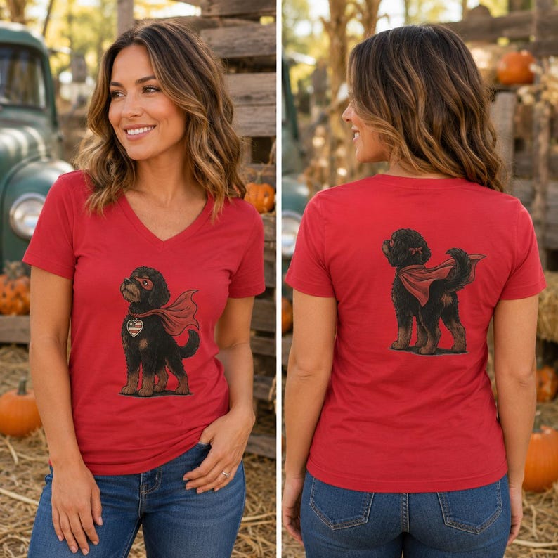 May include: A red V-neck t-shirt featuring a graphic of a black dog wearing a red superhero cape and mask on the front, and the same black dog wearing a red cape on the back. The dog has tan markings on its paws and a small red bow on its head.