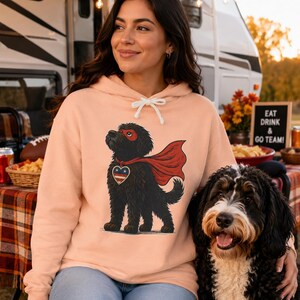 Pu&ograve; includere: Una felpa con cappuccio color pesca che presenta un'illustrazione grafica di un cane nero che indossa un mantello da supereroe rosso, una maschera rossa e un emblema a forma di cuore con stelle e strisce rosse, bianche e blu.