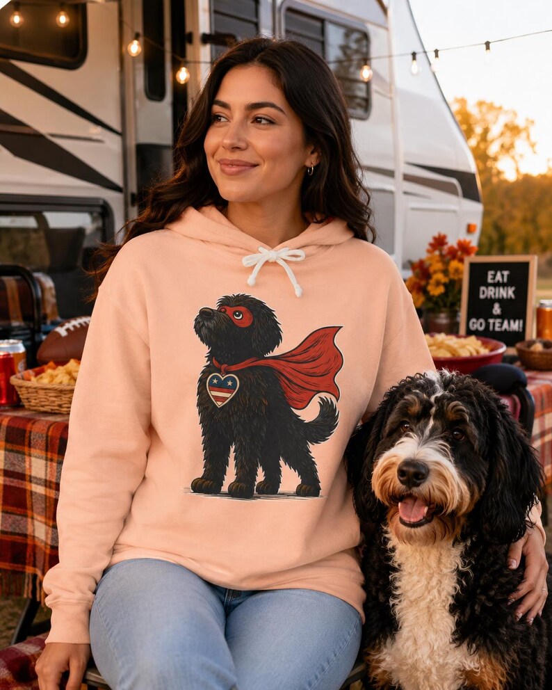 Pu&ograve; includere: Una felpa con cappuccio color pesca che presenta un'illustrazione grafica di un cane nero che indossa un mantello da supereroe rosso, una maschera rossa e un emblema a forma di cuore con stelle e strisce rosse, bianche e blu.